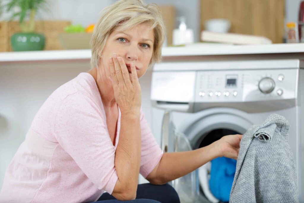 A woman looks thoughtful while loading a sweater into a washing machine in a laundry room.