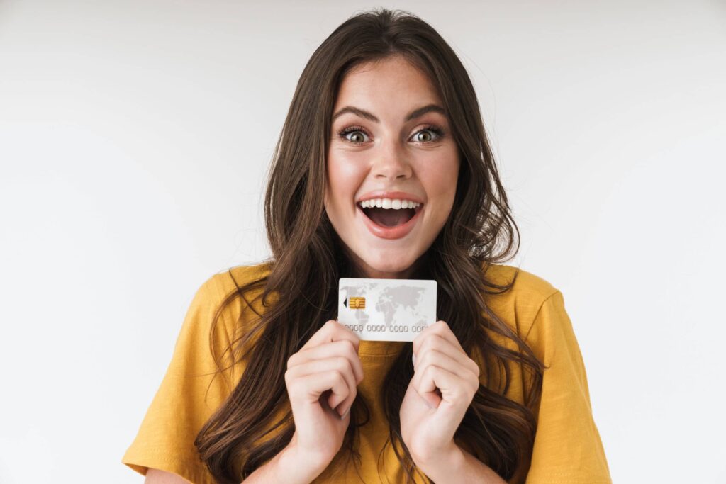 A woman in a yellow shirt holds a credit card, smiling with an excited expression against a plain background, eager to maximize her credit card rewards.
