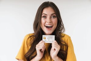 A woman in a yellow shirt holds a credit card, smiling with an excited expression against a plain background, eager to maximize her credit card rewards.