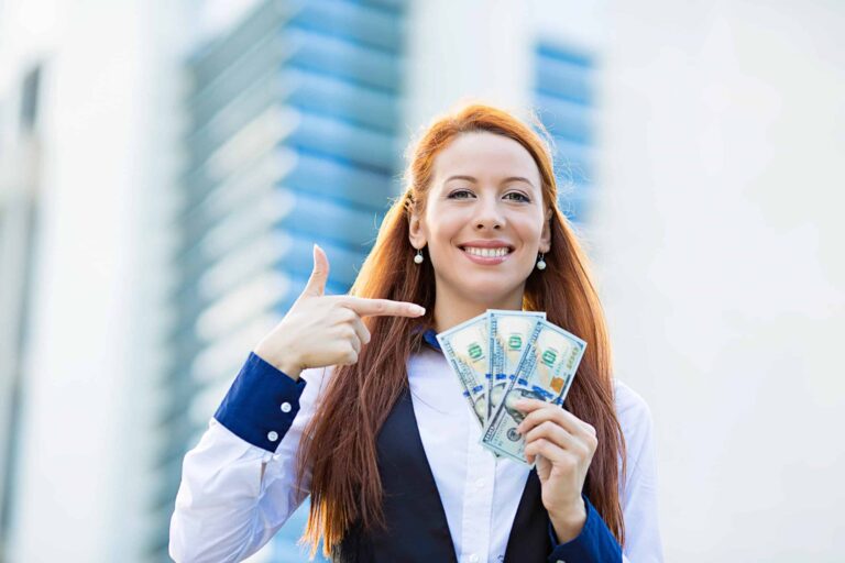 A woman with long red hair holds several US hundred-dollar bills in one hand and points to them with the other, smiling. She is wearing a white shirt and black vest.