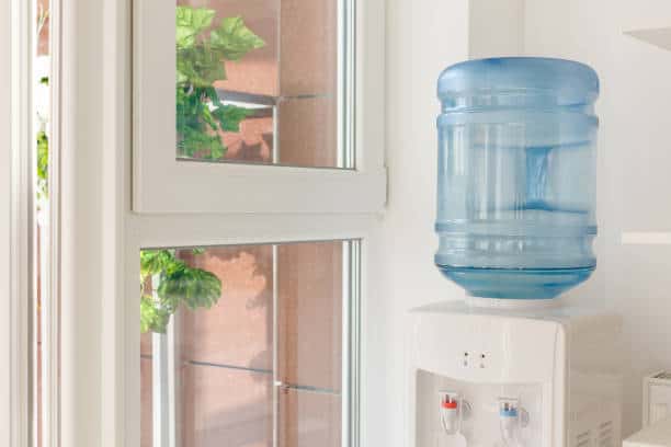 Large water cooler with a blue jug beside a window, with greenery visible outside.