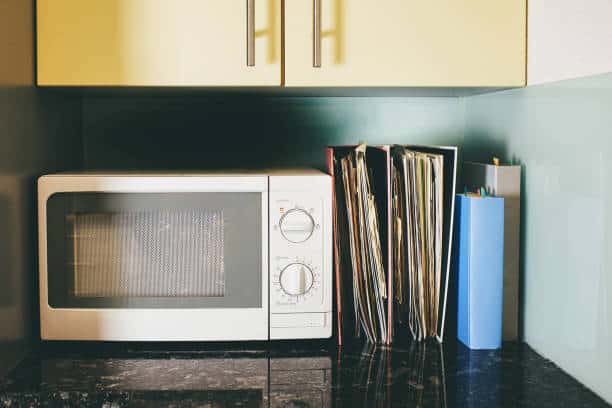 Microwave on a kitchen counter next to a stack of vinyl records and a blue container with kitchen utensils.