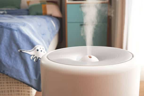 Close-up of a humidifier releasing mist in a bedroom with a dinosaur-themed bedspread and a dresser in the background.