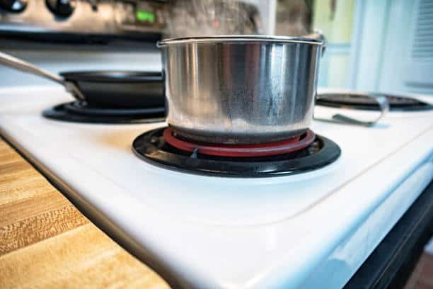 A stainless steel pot with steam rising sits on an electric stove burner. Nearby, a frying pan is on another burner. The stove and countertop are white with wood accents.