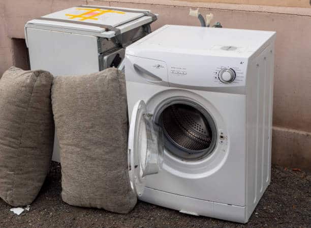 Abandoned washing machine with open door and broken dishwasher on the street, alongside two large cushions.