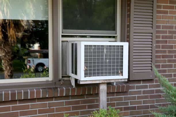 A window air conditioning unit is installed in a brick wall, supported by a wooden brace.