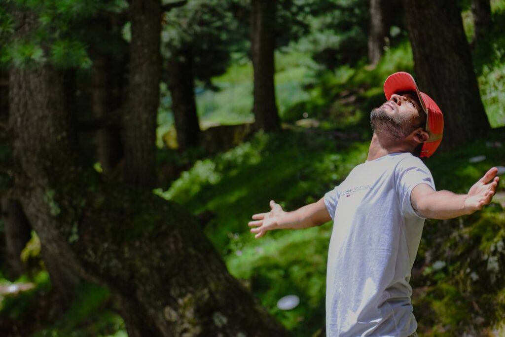 Man in a red cap and white t-shirt stands outdoors with arms outstretched, looking up. Surrounded by trees and greenery.