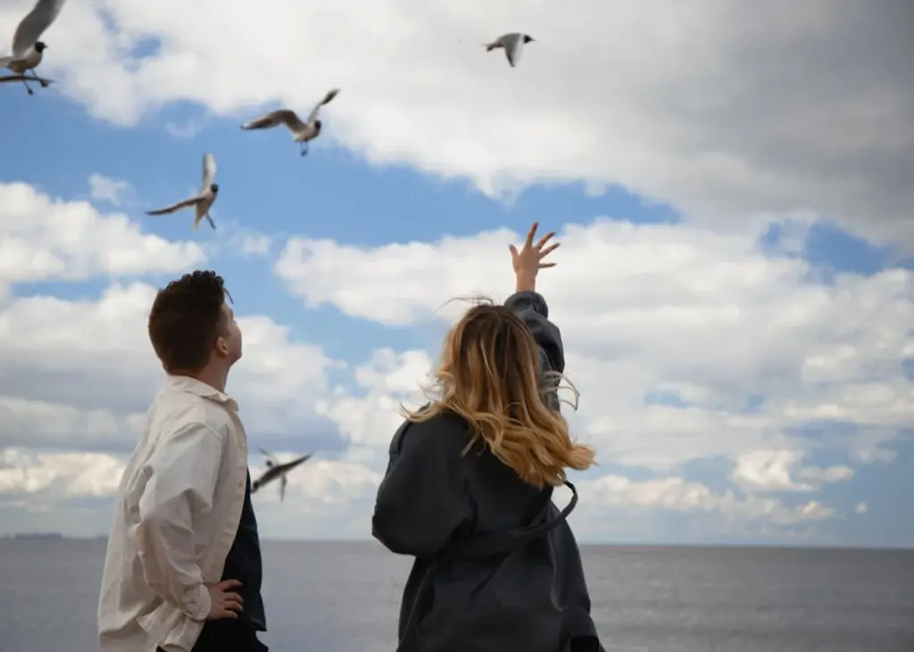 A man and woman stand by the sea, watching and reaching towards seagulls flying in the cloudy sky.
