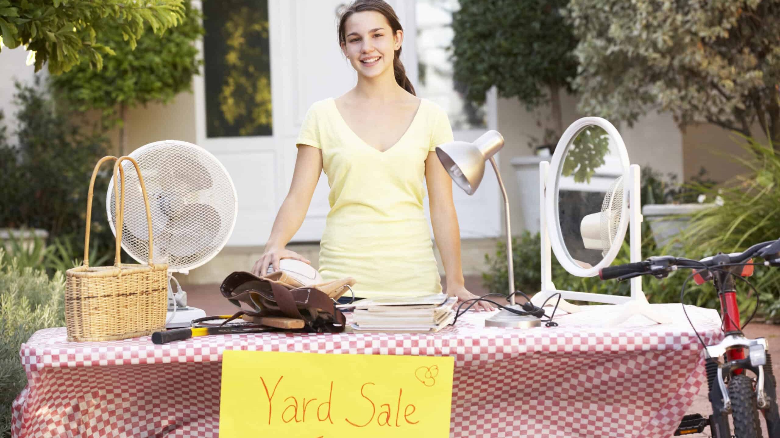 Person standing behind a table with various items and a "Yard Sale Today" sign on it.