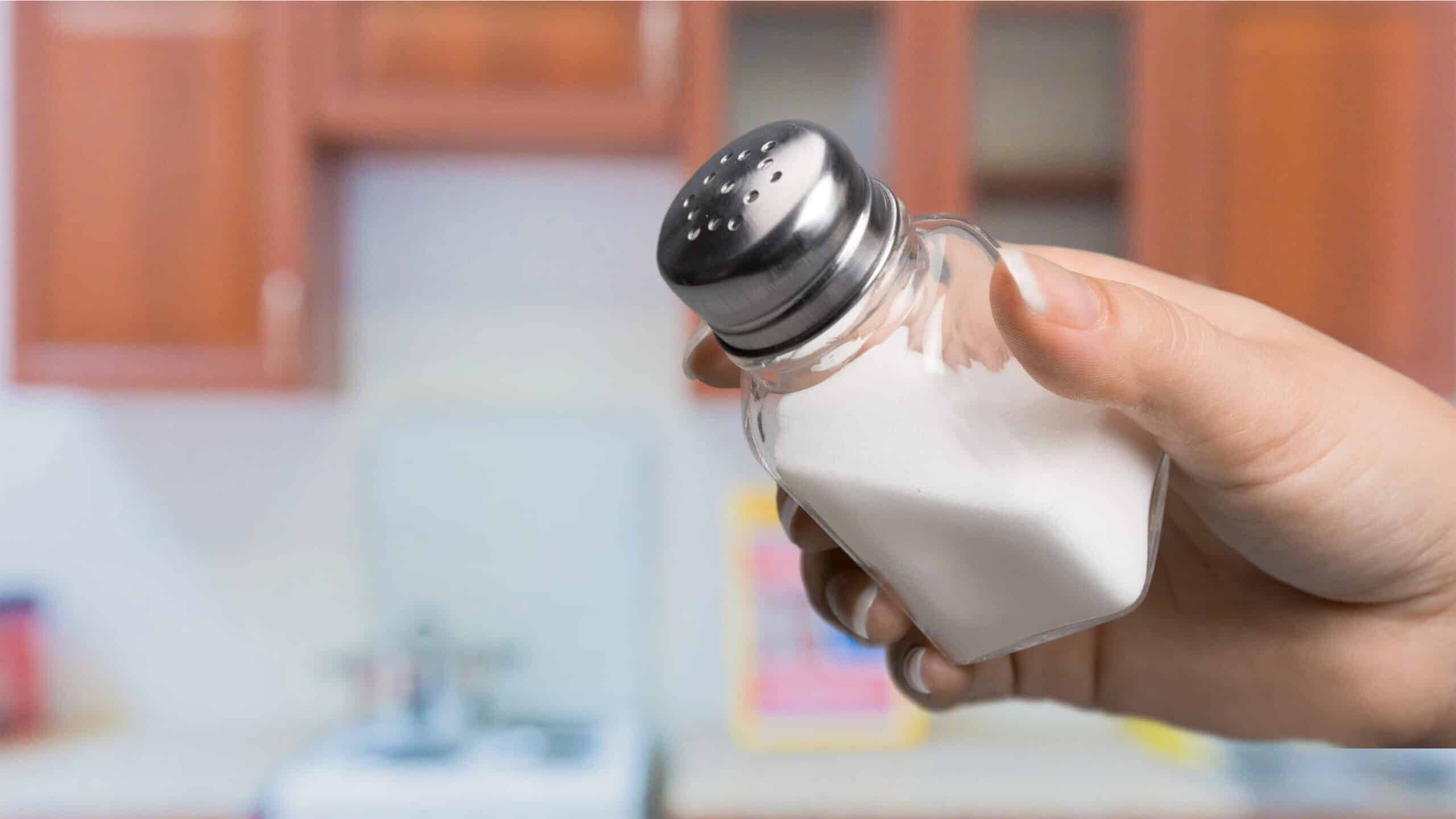 A hand holding a salt shaker in a kitchen setting with wooden cabinets and a stove in the background.