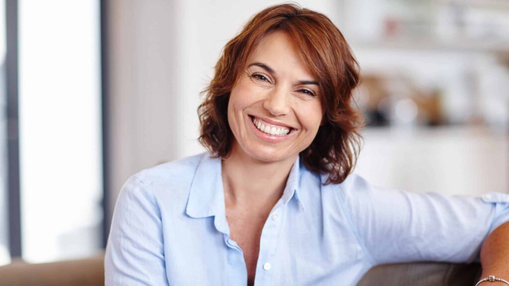 A woman with short brown hair smiles while seated on a sofa, wearing a light blue shirt. The background is softly blurred, suggesting an indoor setting.