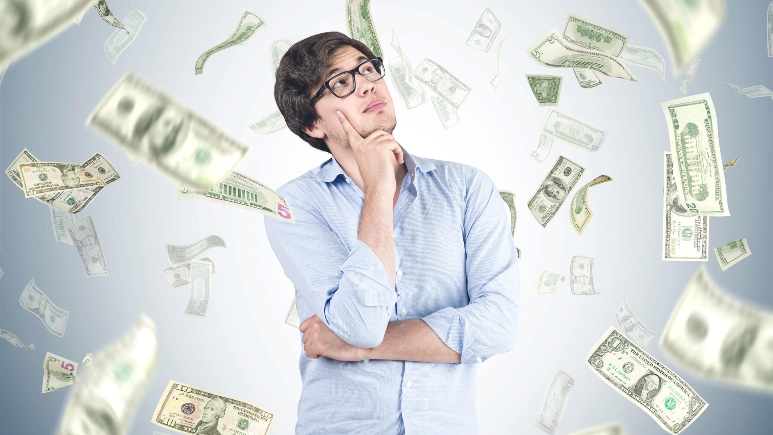 A man in a blue shirt and glasses is looking up thoughtfully, surrounded by falling U.S. dollar bills against a gray background, representing upper class earnings.